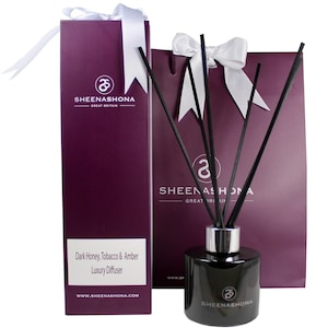 Puede incluir: Una botella difusora de vidrio negro con una tapa plateada y cañas negras. La botella está etiquetada con "Dark Honey, Tobacco & Amber Luxury Diffuser" y "Sheenashona Great Britain". La botella está empaquetada en una bolsa de regalo morada con una cinta blanca.
