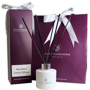 Puede incluir: Una botella blanca con una tapa plateada y palitos difusores negros. La botella está etiquetada con "Very Berry Luxury Diffuser" y "Sheenashona". La botella está empaquetada en una bolsa de regalo morada con una cinta blanca y una caja blanca con una cinta blanca.