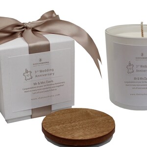 Puede incluir: Una vela blanca en un tarro de vidrio con tapa de madera. La vela está decorada con una etiqueta que dice "3rd Wedding Anniversary" y "Mr & Mrs Davis". La vela está empaquetada en una caja blanca con una cinta de satén marrón.
