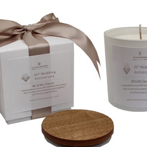 Puede incluir: Una vela blanca con una tapa de madera en una caja de regalo blanca con una cinta de satén beige. La vela tiene una etiqueta que dice "60º Aniversario de Boda Sr. y Sra. Dance Felicitaciones por su 60º Aniversario de Boda Aquí para muchos años más maravillosos juntos Mucho amor Louise & Neil xx www.sheenashona.com"