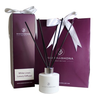 Puede incluir: Una botella blanca de difusor de lujo White Linen con varillas difusoras negras, una bolsa de regalo morada con un lazo blanco y una caja morada con un lazo blanco y el texto "Sheenashona Great Britain" y "www.sheenashona.com".