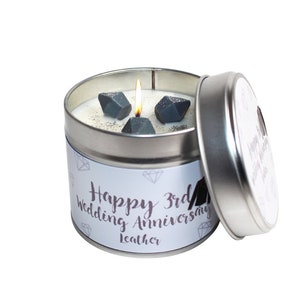 Puede incluir: Una vela en una lata plateada con una etiqueta blanca que dice "Happy 3rd Wedding Anniversary Leather". La vela tiene una mecha encendida y tres decoraciones grises en forma de diamante en la parte superior.