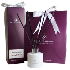 Puede incluir: Una botella blanca de difusor de lujo Amber Gold con varillas difusoras negras y una bolsa de regalo blanca con un lazo blanco. La botella y la bolsa tienen el logotipo de Sheenashoma y el texto "Gran Bretaña".