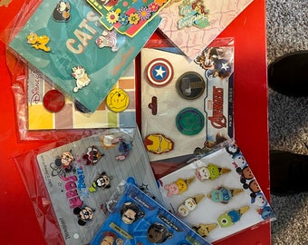 May include: A collection of Disney and Marvel character pins, including characters from The Incredibles, Cats, Captain America, Avengers, and Stitch. The pins are displayed on colorful cards with the Disney and Marvel logos.
