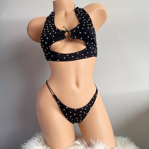 May include: Black two-piece halter top and thong bikini set with white polka dots. The halter top has a keyhole cutout and tie detail. The thong has thin straps.