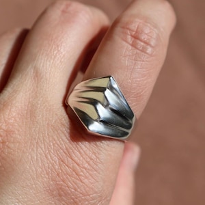 May include: A silver ring with a geometric design. The ring features a rectangular face with a ridged texture, reflecting light. The ring is worn on a finger, showcasing its polished surface and modern aesthetic.
