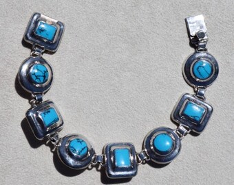 May include: Silver-toned bracelet featuring alternating square and round turquoise-colored stones. The bracelet has a clasp closure and is composed of multiple connected segments. The turquoise stones are set in a silver metal setting.