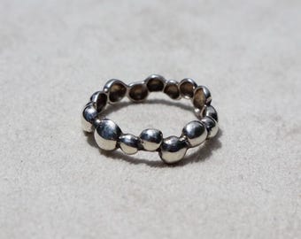 May include: A silver ring featuring a band composed of multiple small, smooth, rounded spheres of varying sizes connected together. The metal has a polished, reflective finish, creating a continuous, irregular, and organic circular shape.