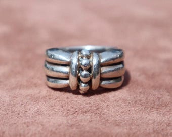 May include: A silver ring with a unique design. The ring features three horizontal bands with a series of small, raised spheres in the center. The ring is set against a muted, reddish-brown background.