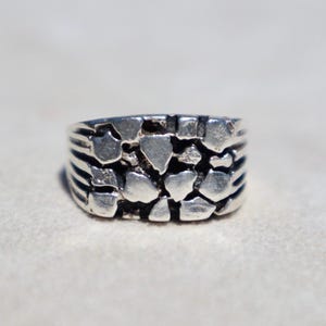 Puede incluir: Un anillo de tono plateado con una superficie texturizada y multifacética compuesta por numerosas formas geométricas pequeñas, irregulares y elevadas. La banda tiene una construcción ancha y sólida con acabado metálico y áreas oscuras profundas.