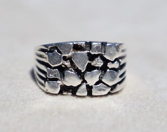 May include: A silver-toned ring featuring a textured, multi-faceted surface composed of numerous small, irregular, raised geometric shapes. The band has a wide, solid construction with a metallic finish and deep, dark recessed areas between the segments.