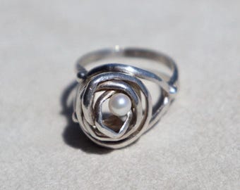 May include: A silver ring featuring a coiled, wire-like band that forms a circular, open-work design. A single, small, round white pearl is set within the center of the silver wire coils, creating a focal point for the metallic, textured band.