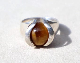 May include: A silver ring featuring a large, oval, polished tiger's eye gemstone. The stone is a warm brown color with natural striations. The ring's setting has a modern, open design, with curved silver elements cradling the stone.