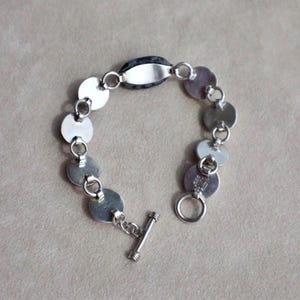 May include: A silver bracelet featuring a series of circular links connected by small rings. The bracelet has a toggle clasp and a central oval-shaped link. The bracelet is made of a shiny, metallic material.