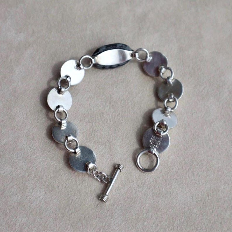 May include: A silver bracelet featuring a series of circular links connected by small rings. The bracelet has a toggle clasp and a central oval-shaped link. The bracelet is made of a shiny, metallic material.