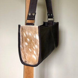 Puede incluir: Un bolso de mano de cuero marrón con una correa larga y una solapa frontal de piel de vaca manchada.