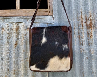 Könnte beinhalten: Eine braune Ledertasche mit einem weißen und braunen Kuhfellmuster. Die Tasche hat einen langen Riemen und einen Klappverschluss.