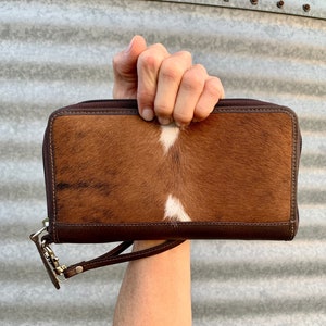 May include: A brown leather wallet with a white and brown cowhide pattern. The wallet has a zipper closure and a wrist strap.