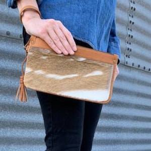 May include: A woman is wearing brown leather boots with buckles and a brown and white cowhide clutch with a tassel.