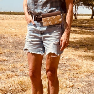 May include: A woman wearing denim shorts and a plaid shirt with a brown and white cowhide fanny pack worn around her waist.