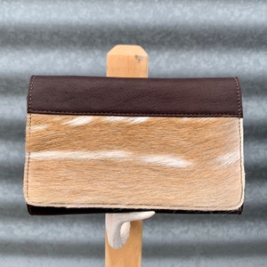 May include: A brown leather wallet with a tan and white fur flap. The wallet is closed and hanging from a wooden stand.