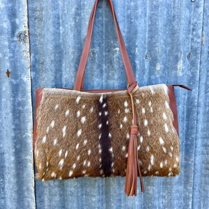 Puede incluir: Un bolso tote de cuero marrón con un panel frontal de piel de venado manchado y un borla de cuero marrón. El bolso tiene dos correas de cuero marrón.