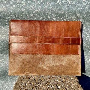 May include: A brown leather clutch with a brown and white cowhide flap. The clutch has a stitched leather strap.