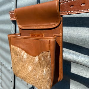 May include: A brown leather belt bag with a flap closure and a front pocket featuring a patch of light brown and white animal hide. The bag is attached to a brown leather belt with white stitching. The text "Silla Texas" is visible.