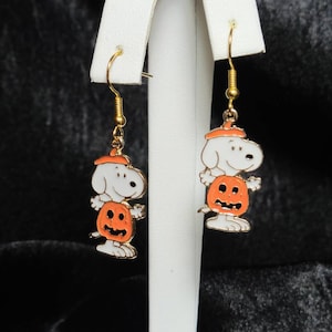 Puede incluir: Pendientes dorados de Halloween. Cada pendiente presenta un personaje de perro de dibujos animados blanco con un sombrero naranja y de pie sobre una calabaza naranja. Los pendientes tienen cierres de gancho dorados y se exhiben sobre un soporte blanco.