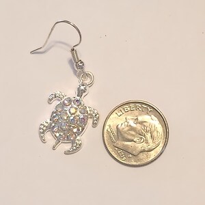 Puede incluir: Pendiente plateado de tortuga marina con cristales iridiscentes. El pendiente se muestra junto a una moneda para comparar el tamaño. La tortuga tiene un gancho para orejas perforadas.