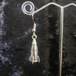 Può includere: Un orecchino a forma di razzo color argento con un piccolo gancio e un dettaglio a molla elicoidale. Il razzo ha piccole alette e un design dettagliato. L'orecchino è esposto su un supporto in metallo argentato su sfondo scuro.