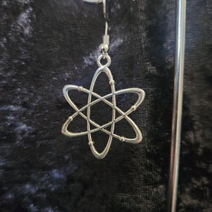 Könnte beinhalten: Silberfarbene Atom-Ohrringe mit Hakenverschluss. Die Ohrringe zeigen ein stilisiertes Atomdesign mit kreisenden Ringen und kleinen Kugeln vor dunklem Hintergrund. Die Ohrringe sind leicht und für den täglichen Gebrauch geeignet.