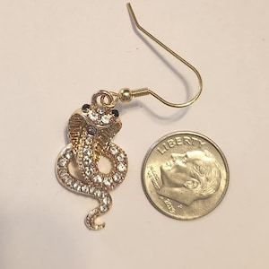 May include: Gold-coloured cobra-shaped earring with clear crystal accents. The snake's head and body are adorned with sparkling crystals, and the eyes are black. The earring has a gold-coloured fish hook.