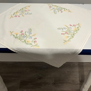 Pu&ograve; includere: Tovaglia bianca con ricami floreali in tonalit&agrave; rosa, blu, giallo e verde. La tovaglia &egrave; su un tavolo bianco con un bordo blu scuro.