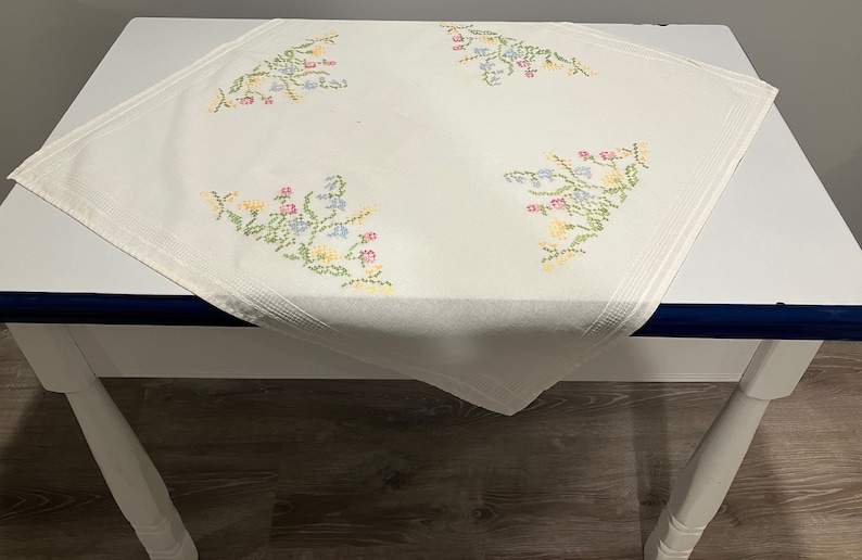 Pu&ograve; includere: Tovaglia bianca con ricami floreali in tonalit&agrave; rosa, blu, giallo e verde. La tovaglia &egrave; su un tavolo bianco con un bordo blu scuro.