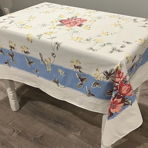 May include: A white rectangular tablecloth with a floral pattern. The design features red and yellow flowers with green stems and leaves. A light blue border with a scalloped edge runs along the edges of the tablecloth.