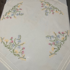 Pu&ograve; includere: Tovaglia bianca con un delicato ricamo floreale in tonalit&agrave; di rosa, blu, giallo e verde. Il ricamo presenta piccoli fiori e foglie disposti in un motivo simmetrico.