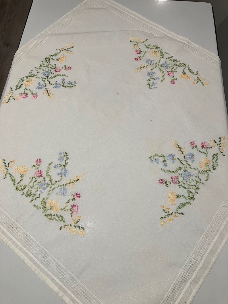 Pu&ograve; includere: Tovaglia bianca con un delicato ricamo floreale in tonalit&agrave; di rosa, blu, giallo e verde. Il ricamo presenta piccoli fiori e foglie disposti in un motivo simmetrico.