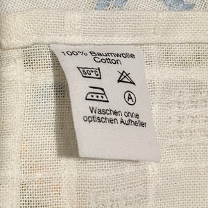 Pu&ograve; includere: Etichetta di cura del tessuto bianca con testo nero e simboli. Il testo recita "100% Baumwolle Cotton", "60&deg;C", "Waschen ohne optischen Aufheller". I simboli includono un'icona di lavatrice, un triangolo con una X all'interno e un ferro da stiro con tre punti.