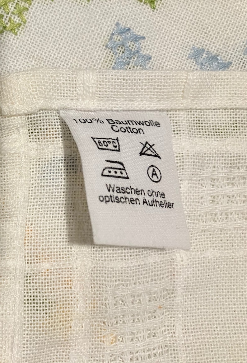 Pu&ograve; includere: Etichetta di cura del tessuto bianca con testo nero e simboli. Il testo recita "100% Baumwolle Cotton", "60&deg;C", "Waschen ohne optischen Aufheller". I simboli includono un'icona di lavatrice, un triangolo con una X all'interno e un ferro da stiro con tre punti.