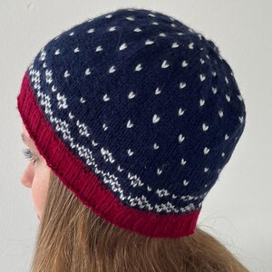 Könnte beinhalten: Eine marineblaue Strickmütze mit rotem Rand und weißen herzförmigen Mustern. Die Mütze hat ein detailliertes Strickdesign und ist für kaltes Wetter geeignet. Die Mütze ist ein Winteraccessoire.