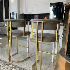 Könnte beinhalten: Zwei goldene Barhocker mit grauen, gepolsterten Sitzen. Die Hocker haben ein schlichtes, modernes Design mit einem goldenen Metallrahmen und einer abgerundeten Basis.