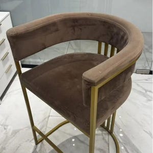 May include: A brown velvet upholstered bar stool with a curved back and armrests. The stool has a gold-colored metal frame with a half-circle base. The seat and back are a solid brown color.