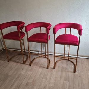 Könnte beinhalten: Drei rote Samt-Barhocker mit goldenen Metallrahmen. Die Hocker haben ein schlichtes, modernes Design mit einer abgerundeten Rückenlehne und einer Fußstütze.