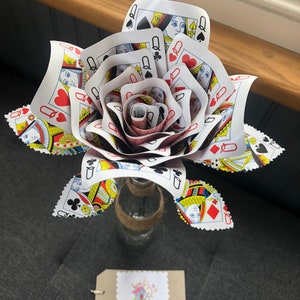 May include: A red, white and black playing card rose made from Queen of Hearts playing cards. The rose is in a clear glass vase with a brown twine bow. The vase is sitting on a grey surface. A brown tag with white text reads "Sam's House of Craft" and a small house icon.