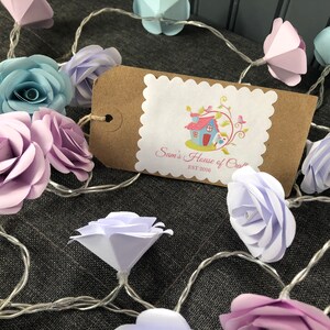 May include: A string of fairy lights with paper flowers in shades of pink, blue, and purple. The flowers are shaped like roses and are strung on a clear wire. A brown tag with the text "Sam's House of Craft Est 2018" is visible in the image.