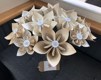 May include: A bouquet of paper flowers made from book pages. Each flower has multiple petals and a white button center. The paper is aged, with visible text. The flowers are arranged against a dark background.