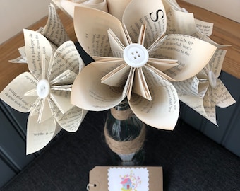 May include: A bouquet of paper flowers made from book pages, with white buttons in the center of each flower. The flowers are arranged in a clear glass bottle with a brown twine bow. A brown tag with the text "Sams House of Craft" is in the foreground.
