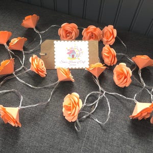 Könnte beinhalten: Eine Lichterkette aus pfirsichfarbenen Papierblumen. Die Blumen sind auf einem durchsichtigen Draht aufgefädelt und mit kleinen weißen Lichtern beleuchtet. Die Lichter liegen auf einer grauen Oberfläche und es befindet sich ein kleines braunes Schild mit dem Text "Sam's House of Craft" darauf.