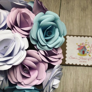 May include: A bouquet of paper flowers in shades of light blue, lavender, and white. The flowers are arranged in a circular shape and have a delicate, layered appearance. The flowers are accented with small, clear beads in the center of each bloom.